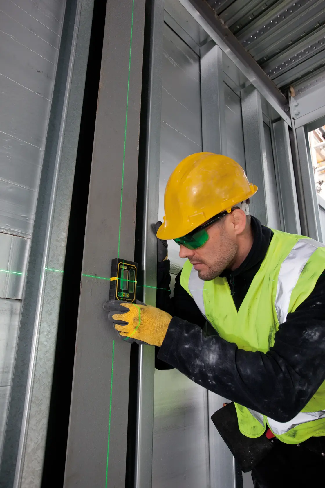 STANLEY FMHT1-74267 Laser-Nivelliergerät im Einsatz an Metallrahmen durch Arbeiter mit gelbem Helm und Weste, Baustelle.