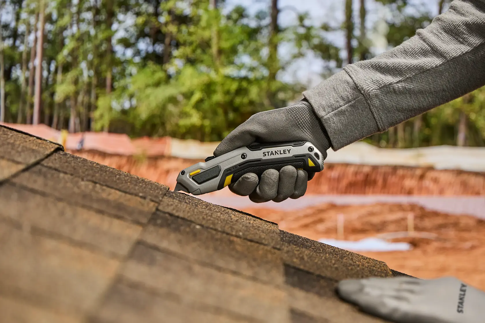Eine behandschuhte Hand verwendet ein Stanley Universalmesser, um Dachschindeln auf einem Dach zu schneiden, mit Bäumen und Baumaterialien im Hintergrund.
