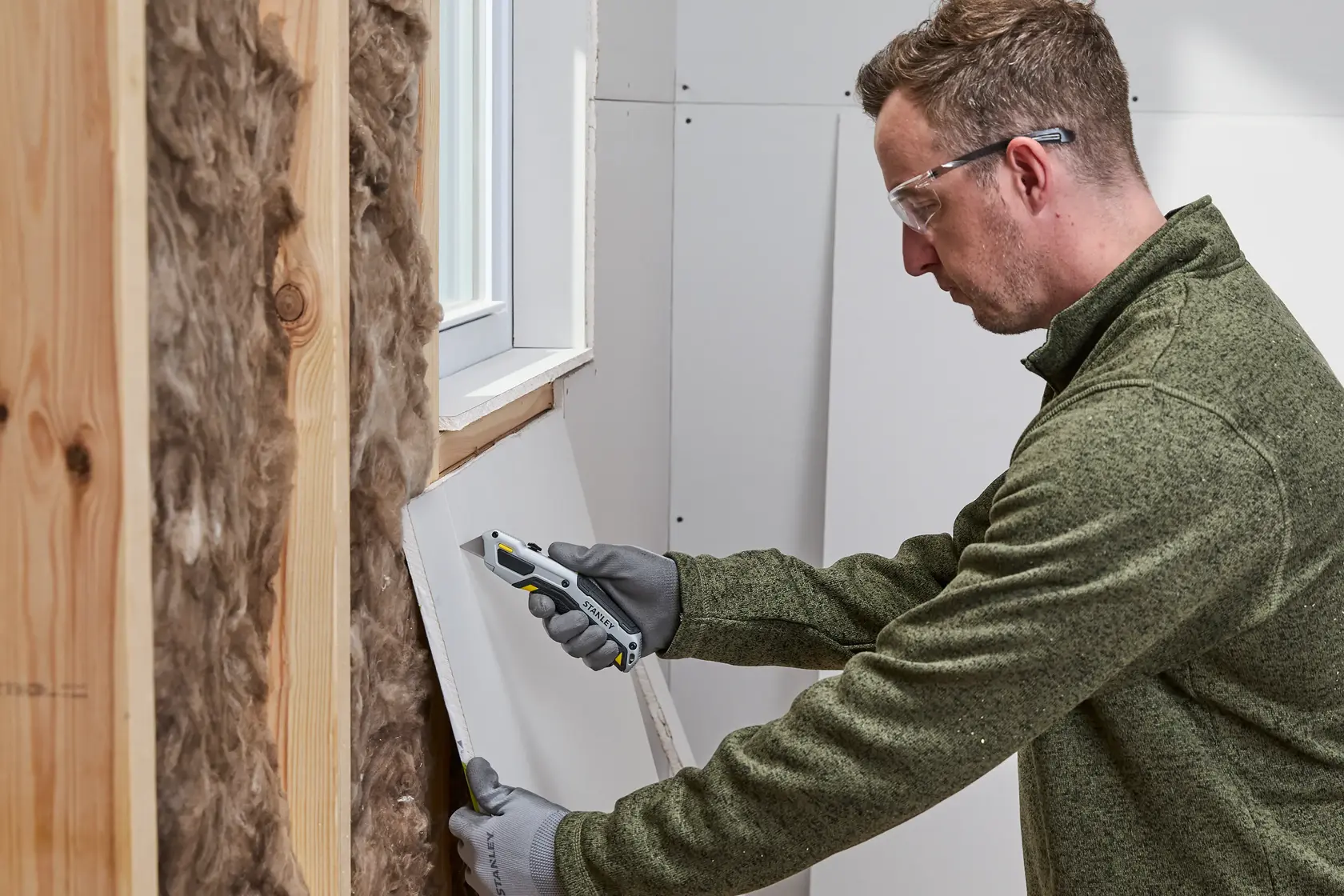 Eine Person mit Handschuhen und grünem Pullover verwendet ein Stanley Universalmesser, um Gipskarton in der Nähe eines Fensters zu schneiden. Die Wand enthält Holzständer und Dämmmaterial.