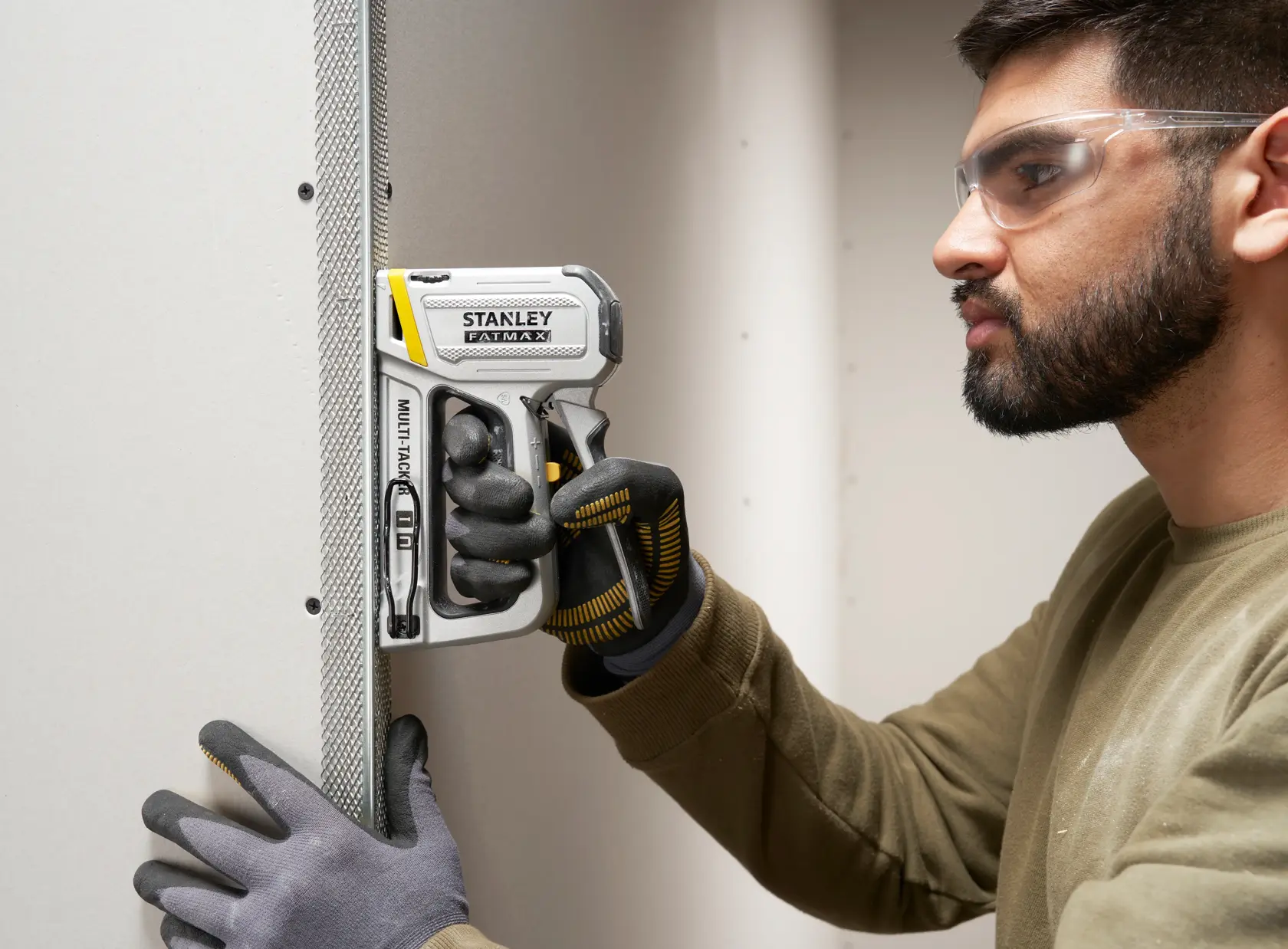 Fotografie des STANLEY FATMAX FMHT70250-0 Tackers im Einsatz an Trockenbauwand, gehalten von behandschuhten Händen.