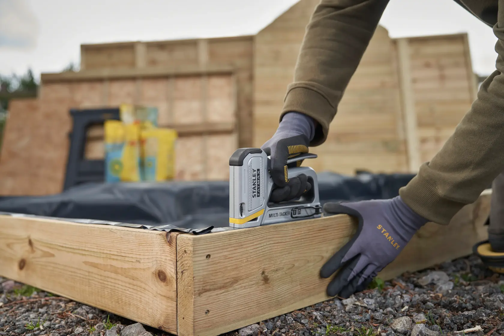 Foto der STANLEY FMHT70250-0 Tacker, der eine schwarze Folie an einem Holzrahmen im Freien befestigt, Person mit STANLEY Handschuhen.