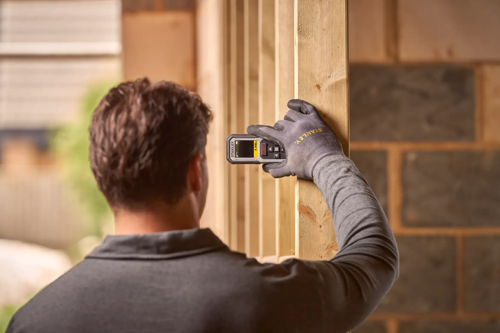 STANLEY FMHT77050-XJ Laser-Entfernungsmesser in behandschuhter Hand an Holzpfosten, Baustellenhintergrund.