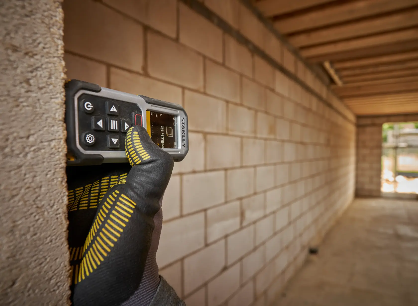 STANLEY FMHT77050-XJ Laser-Entfernungsmesser, gehalten von einer behandschuhten Hand an Ziegelwand auf Baustelle.