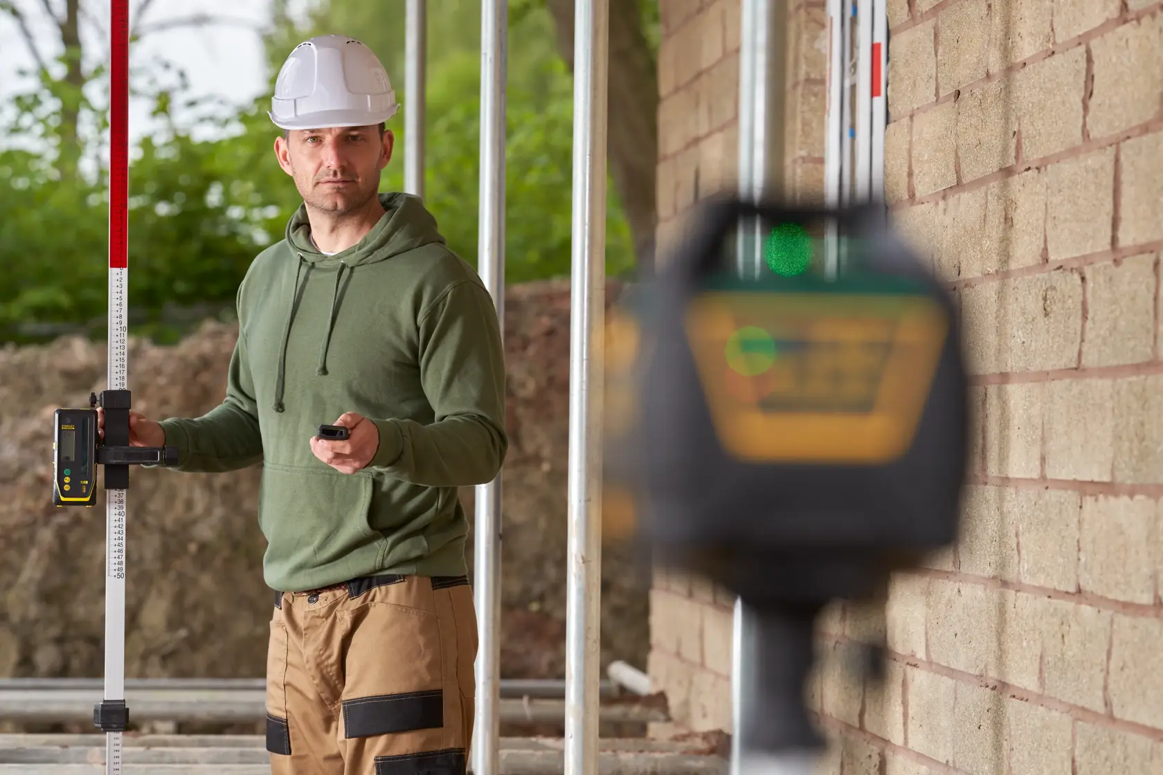 STANLEY FMHT77448 Laser-Nivelliergerät im Einsatz auf einer Baustelle, Arbeiter hält Messlatte und Fernbedienung.