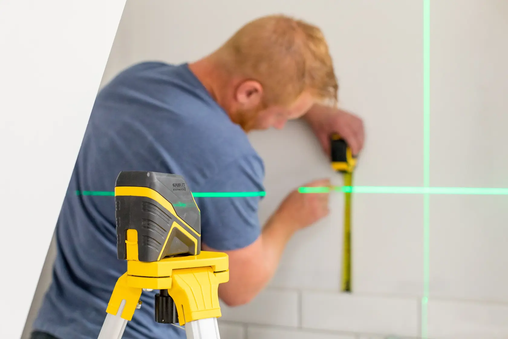 STANLEY FMHT77586-1 Laser-Nivelliergerät auf Stativ projiziert grüne Linien, Person misst Wand.