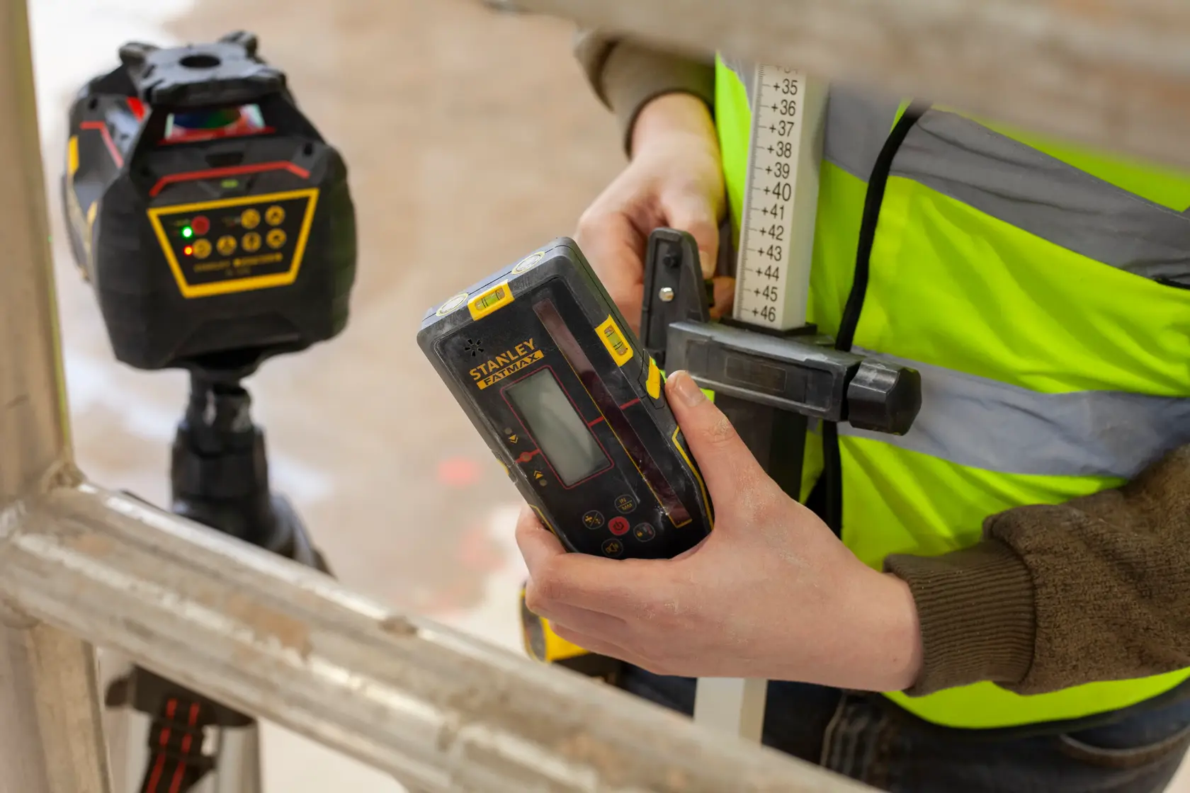 Fotografie des STANLEY FMHT77652-0 Laserdetektors in der Hand, mit Lasergerät im Hintergrund, Baustellenumgebung.