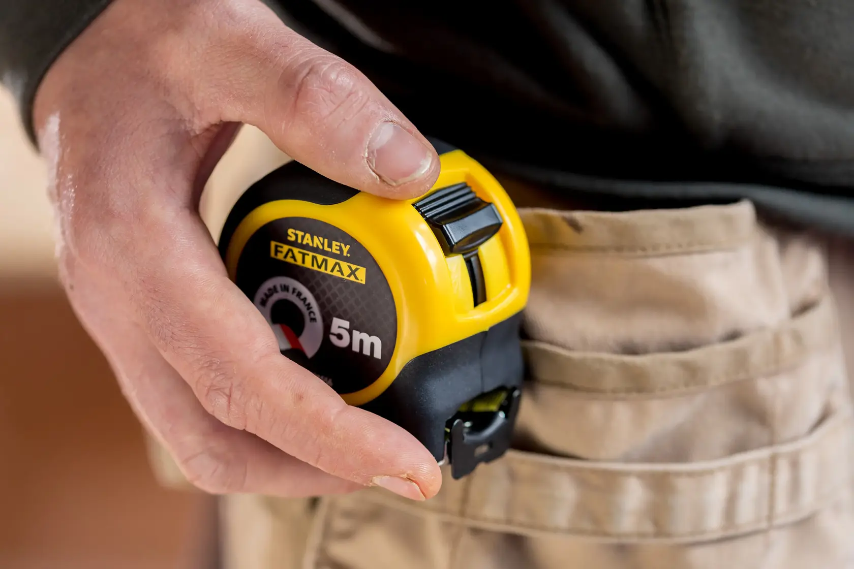 STANLEY FMHT81556-0 Maßband gelb und schwarz, in der Hand neben einer beigen Arbeitshose gehalten.