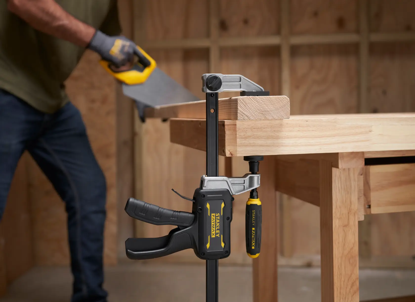 STANLEY FMHT83007-0 Zwinge hält Holz auf Werkbank fest, Nahaufnahme, Person sägt im Hintergrund.