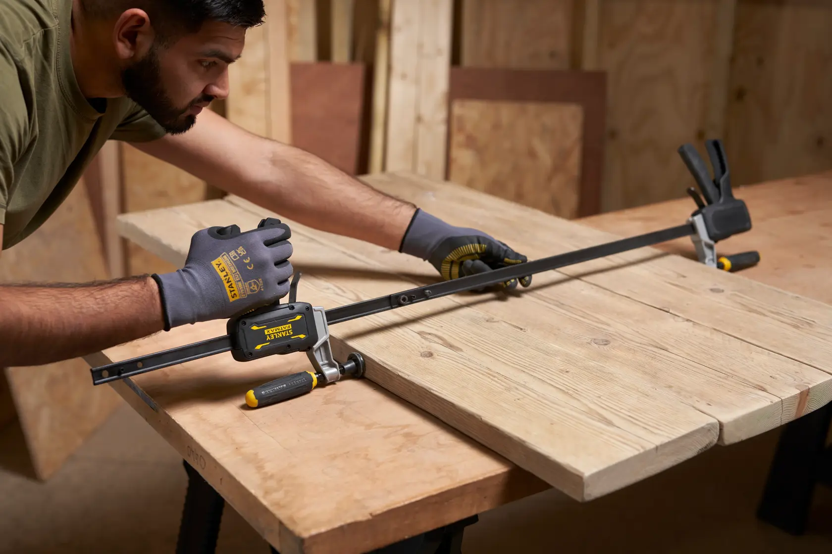 Foto der STANLEY FMHT83008-0 Zwinge, die Holzplatten auf einer Werkbank mit behandschuhter Hand fixiert.