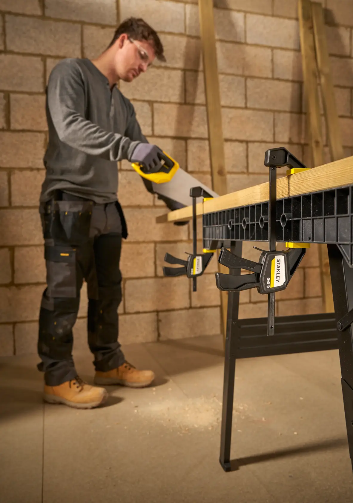 Foto der STANLEY FMHT83271-0 Zwinge, die Holz an der Werkbank in einer Werkstatt sichert, Arbeiter sägt daneben.