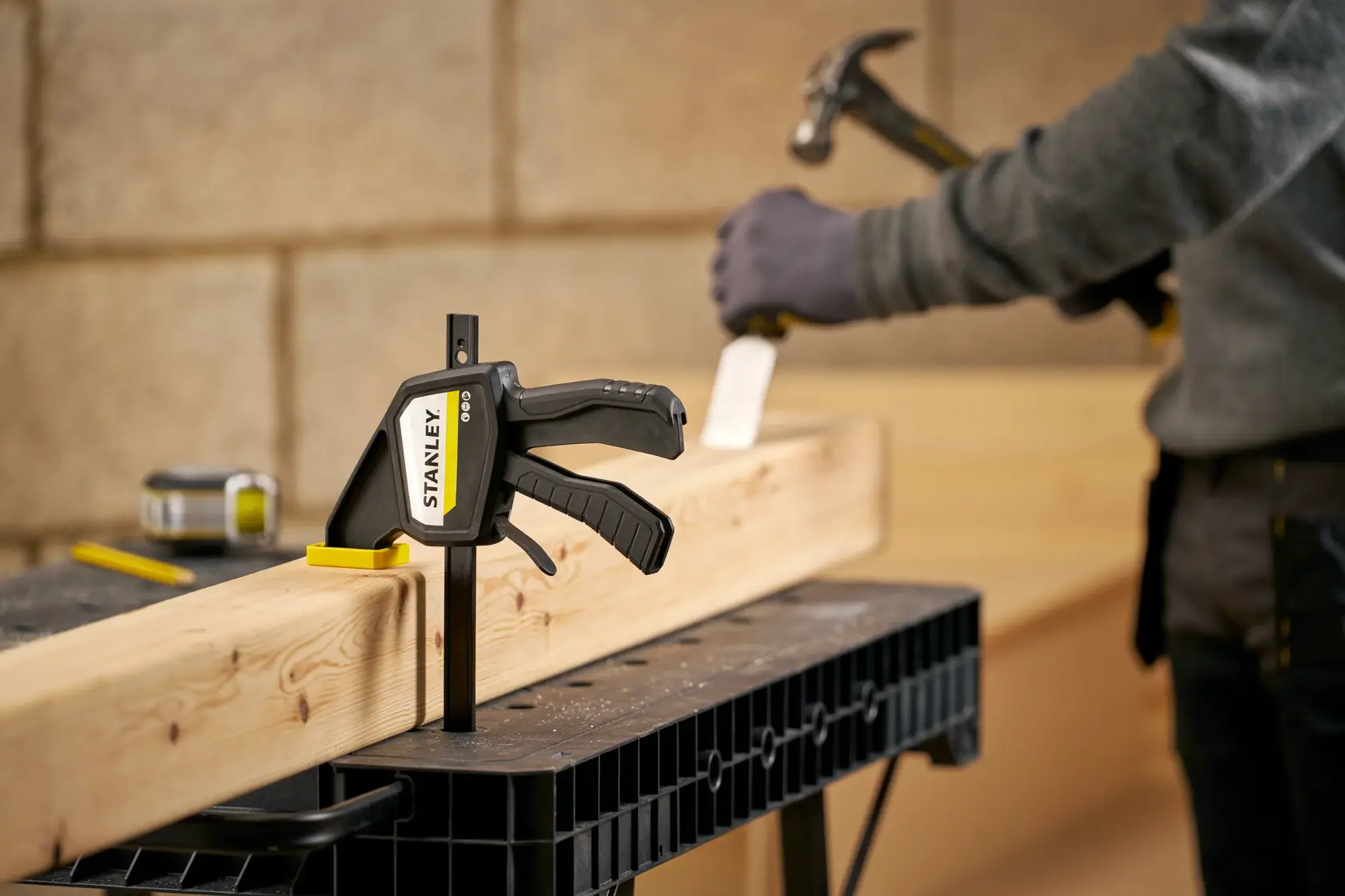 STANLEY FMHT83273-0 Zwinge hält Holzbalken auf Werkbank, Arbeiter mit Hammer im Hintergrund.