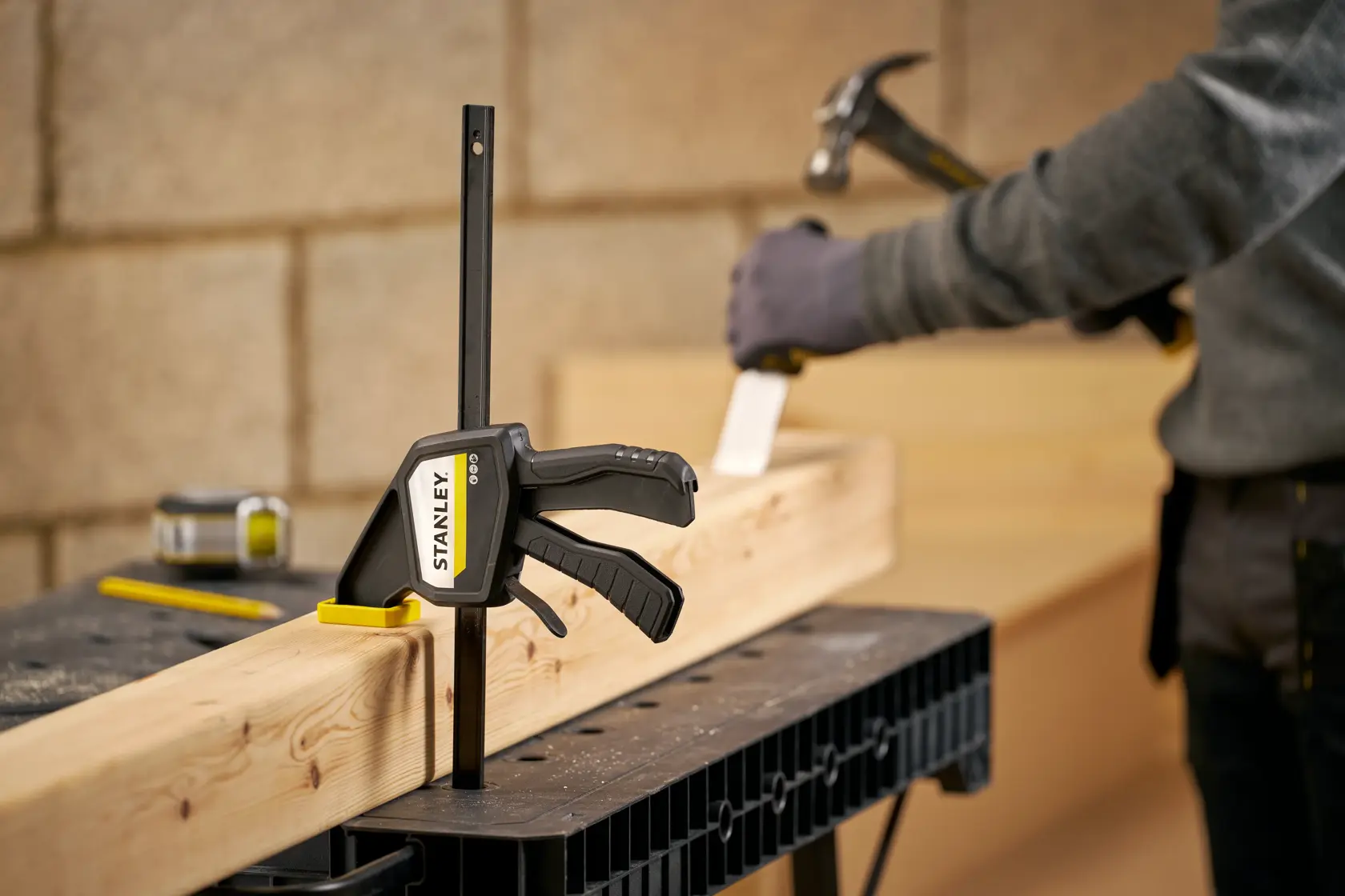 STANLEY FMHT83274-0 Schraubzwinge fixiert Holzbalken auf Werkbank, Person nutzt Hammer im Hintergrund.