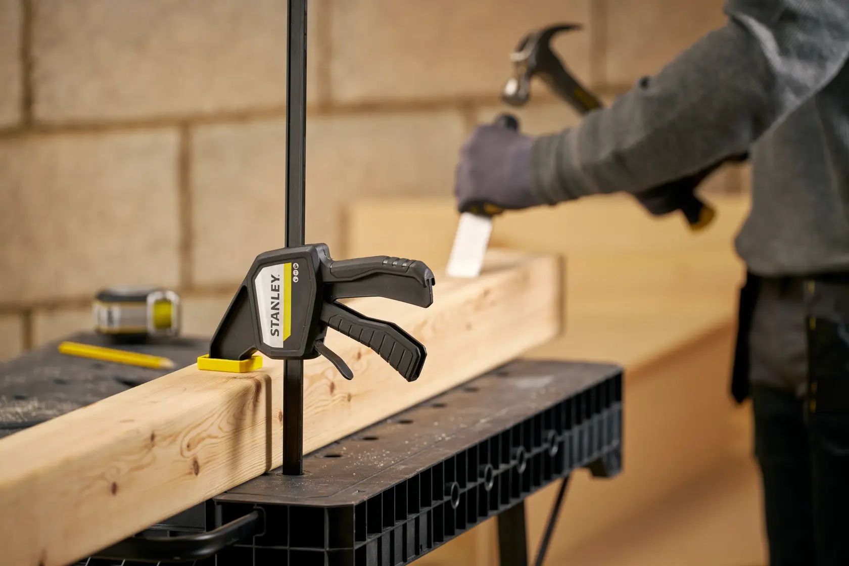 STANLEY FMHT83275-0 Schraubzwinge sichert Holzbalken auf Werkbank, Person mit Hammer im Hintergrund.
