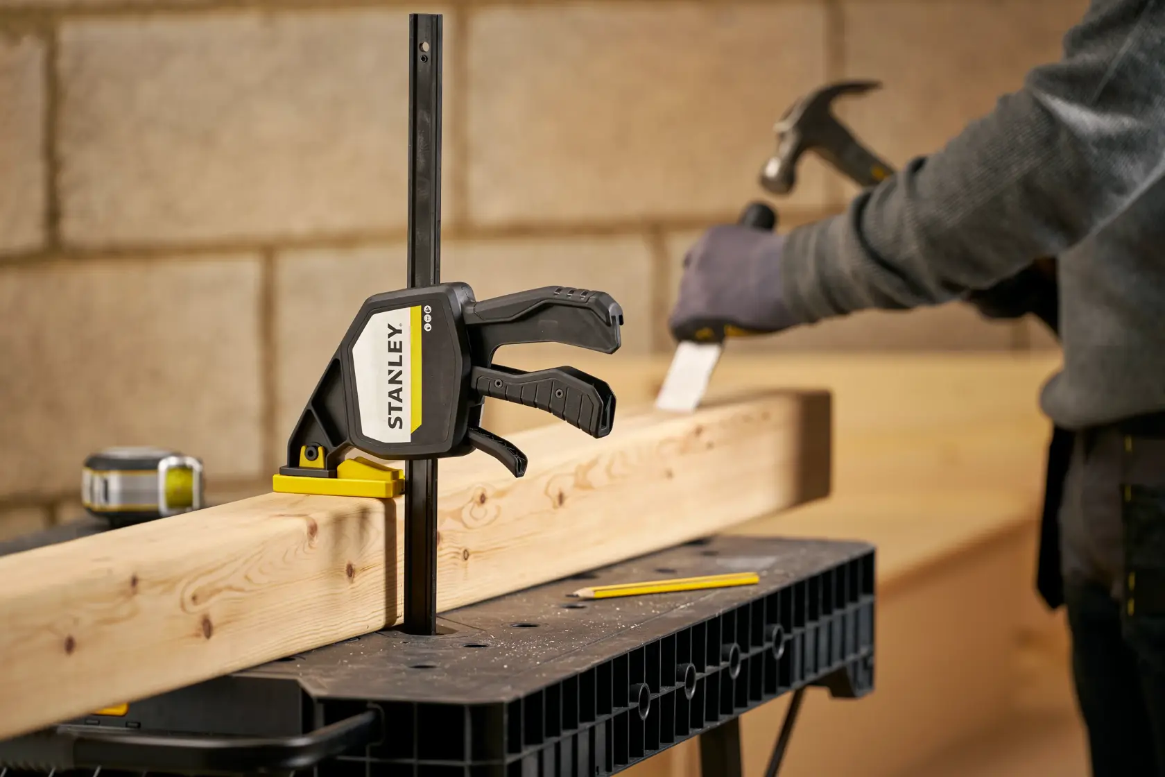 STANLEY FMHT83279-0 Schraubzwinge hält Holz auf Werkbank, Arbeiter mit Hammer im Hintergrund, Werkstattumgebung.