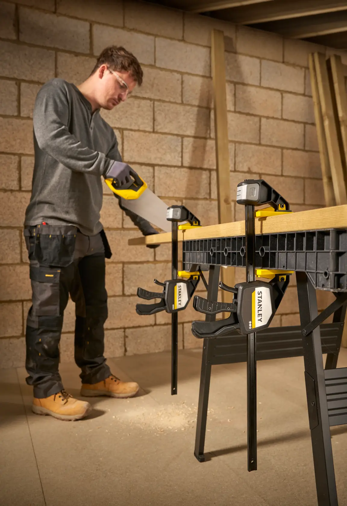 STANLEY FMHT83280-0 Zwinge befestigt Holz auf Werkbank, Person sägt in Werkstatt.