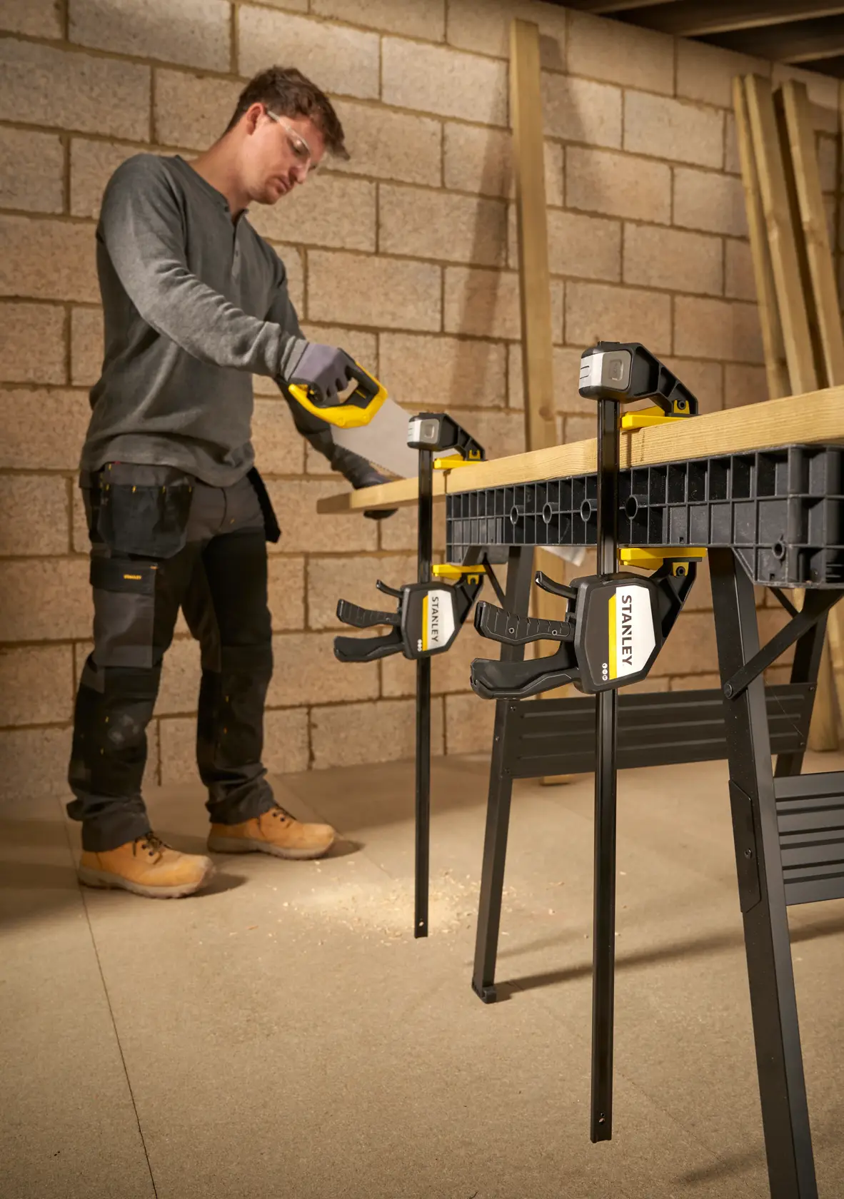 STANLEY FMHT83281-0 Zwinge fixiert Holz auf Werkbank, Person sägt, Werkstattumgebung.