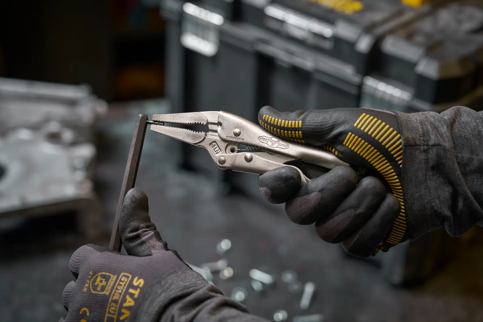 Mit Stanley Feststellzange halten behandschuhte Hände Metall, Teile und Werkzeugkasten im Hintergrund.