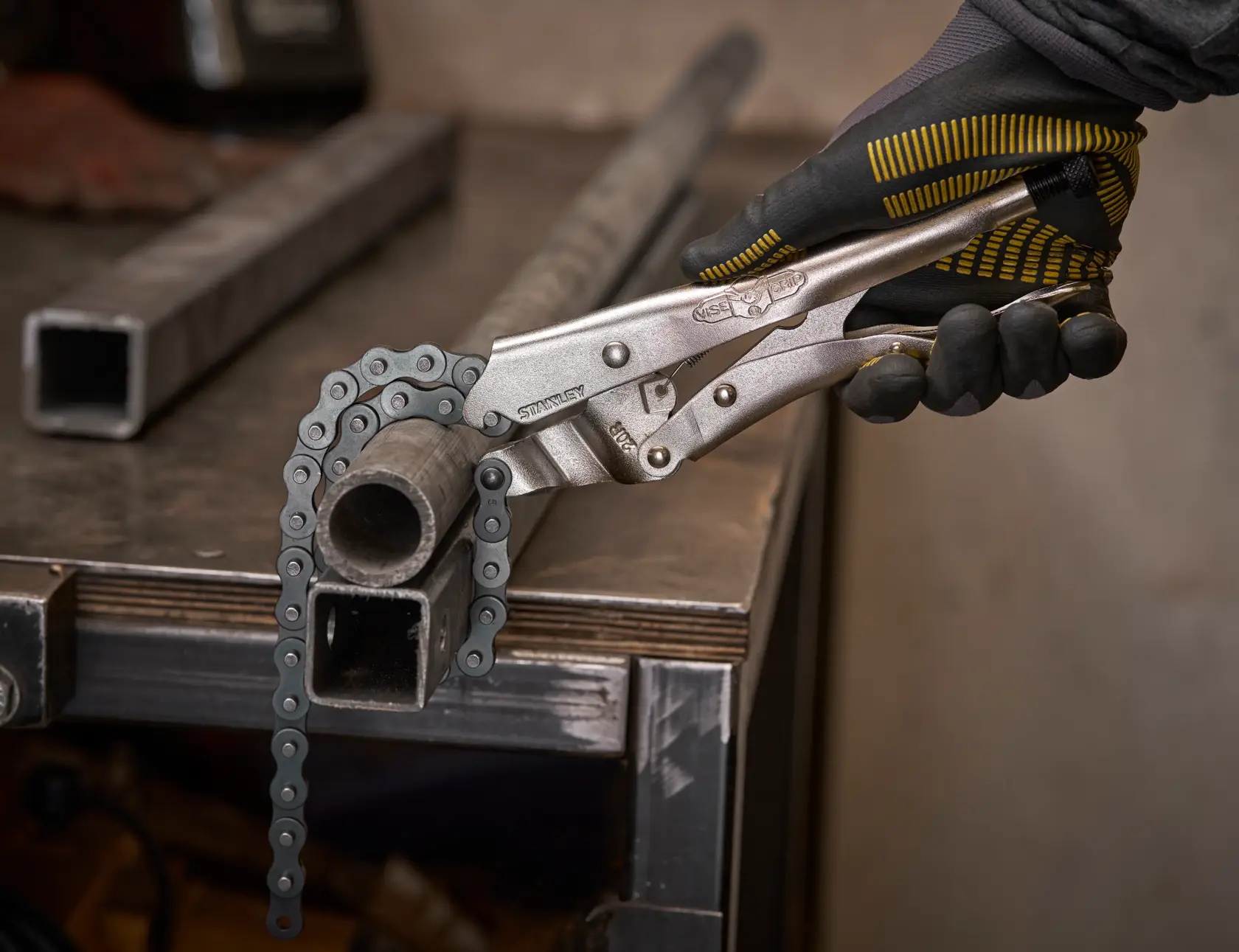 Eine behandschuhte Hand nutzt Stanley Kettenzange, um ein rundes Metallrohr auf einer Werkbank zu greifen.