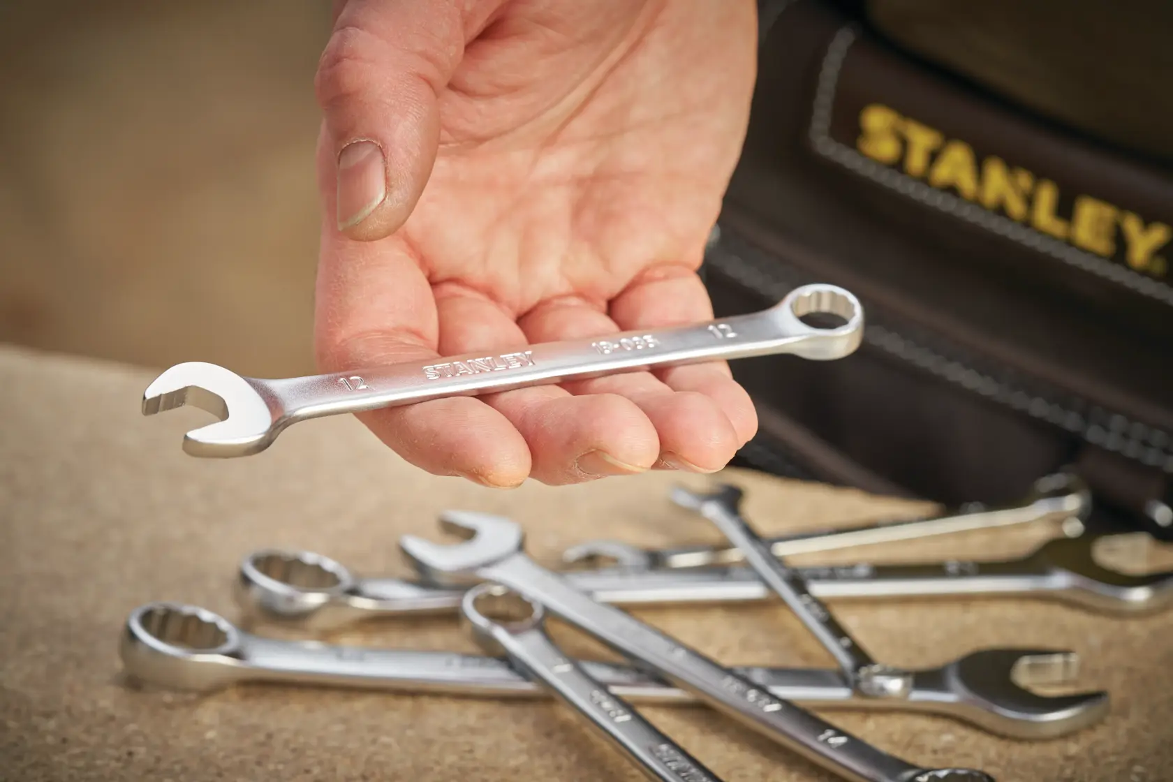 Stanley FMMT13041-0 combination wrench held in hand, with more wrenches on a table, tool belt visible.