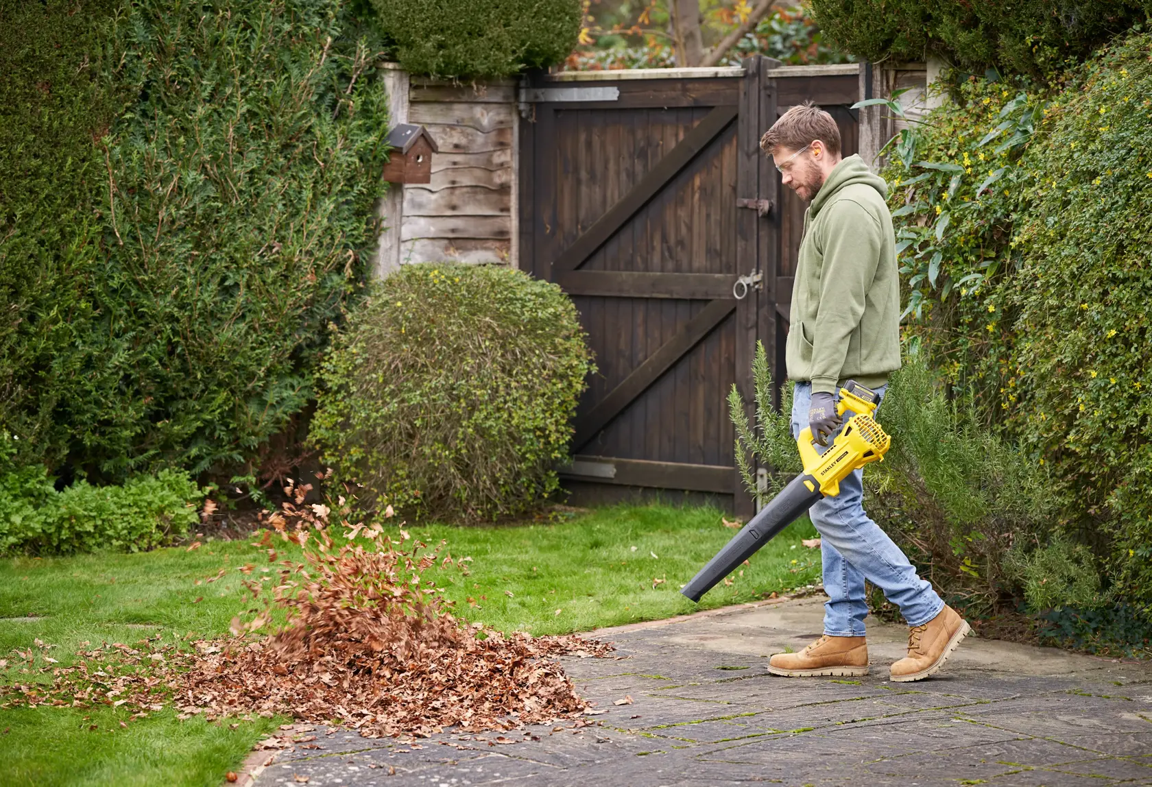 STANLEY SFMCBL714B-XJ Laubbläser im Einsatz, bläst Blätter auf einem Gartenweg, seitliche Außenansicht.