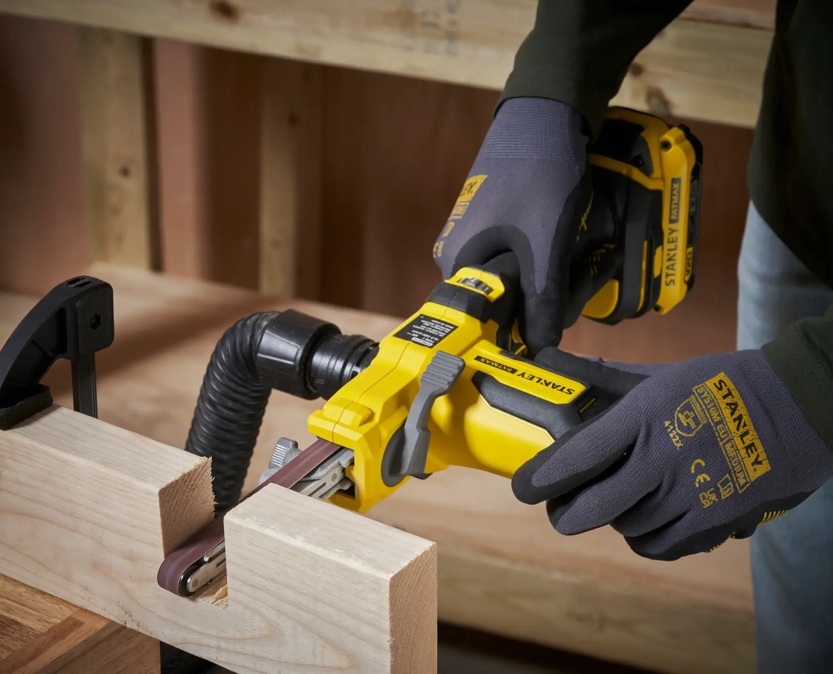 Photo of STANLEY SFMCE210B cordless belt sander in use on a wood surface, worker wearing gloves.