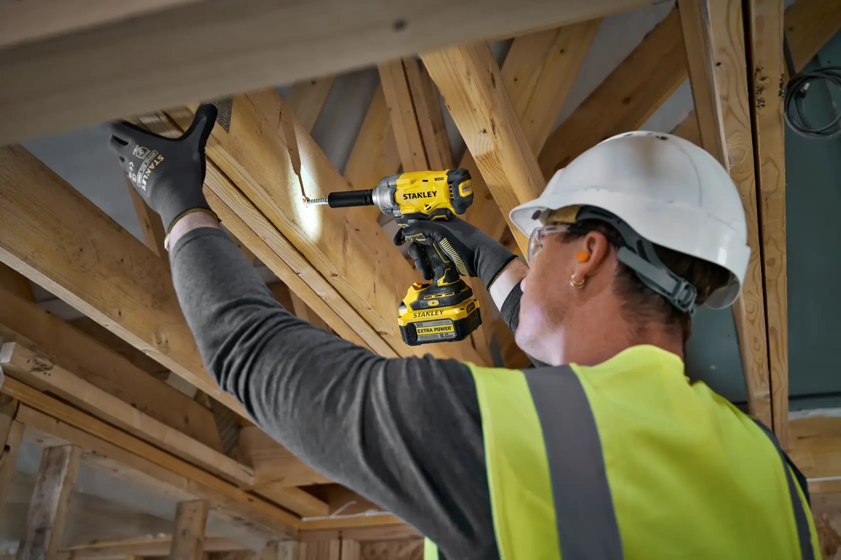 Arbeiter nutzt STANLEY Akku-Bohrmaschine an Holzbalken, mit Handschuhen, Helm und Weste auf Baustelle.