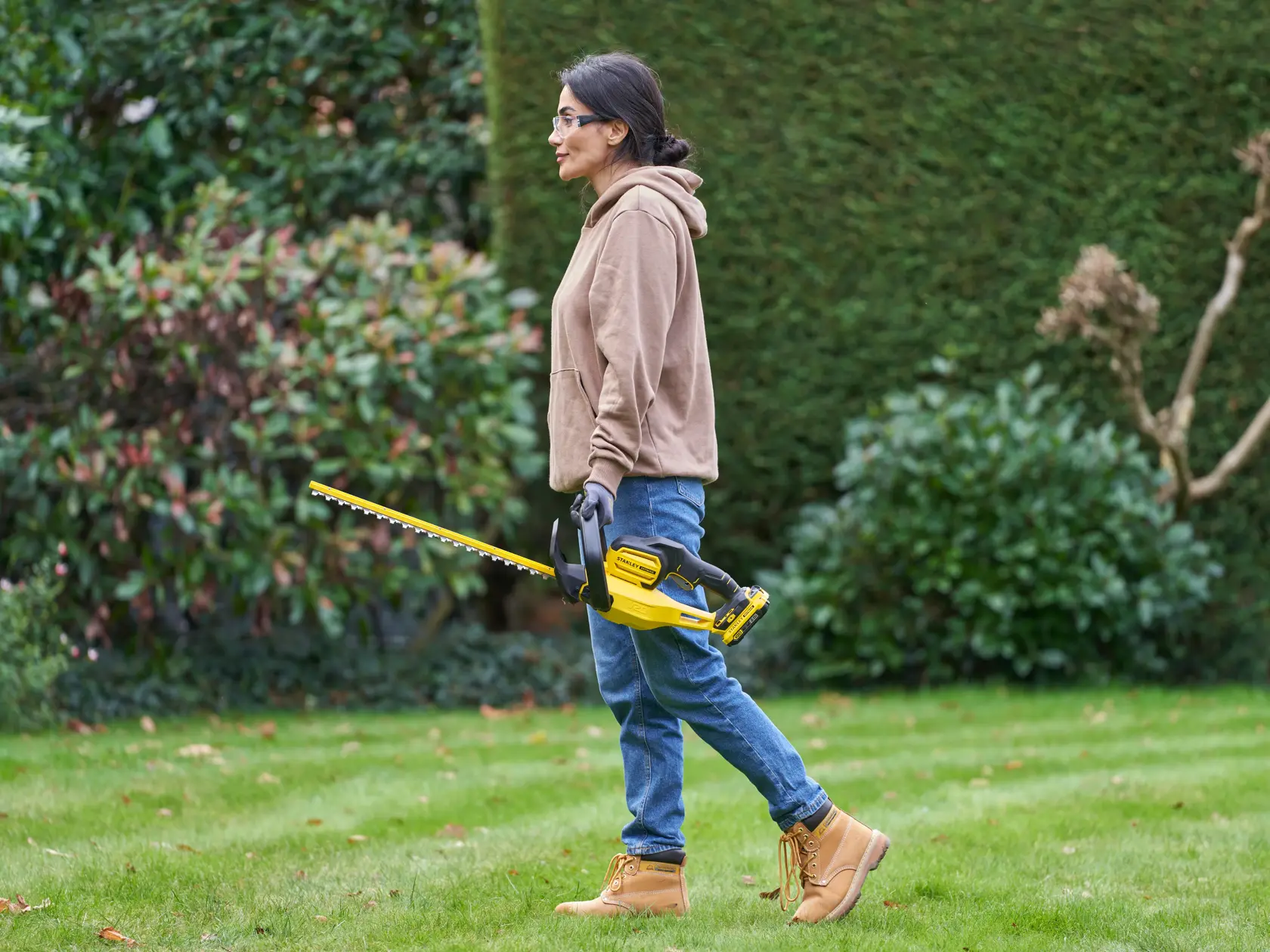 STANLEY SFMCHT650D1 Heckenschneider im Einsatz im Freien, Person hält gelb-schwarzes Gerät nahe grüner Büsche.