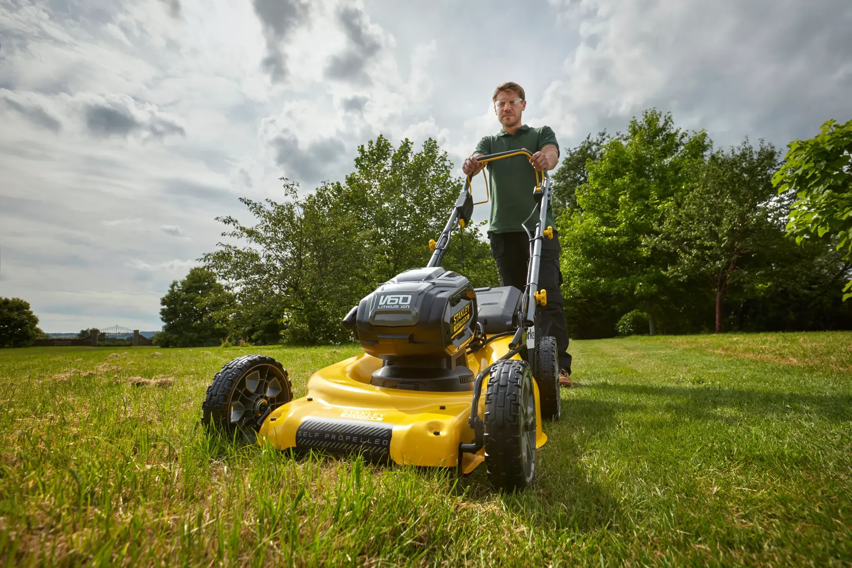 Foto des gelben kabellosen Rasenmähers STANLEY SFMCMS2653B in Betrieb auf Gras, Vorderansicht im Freien.