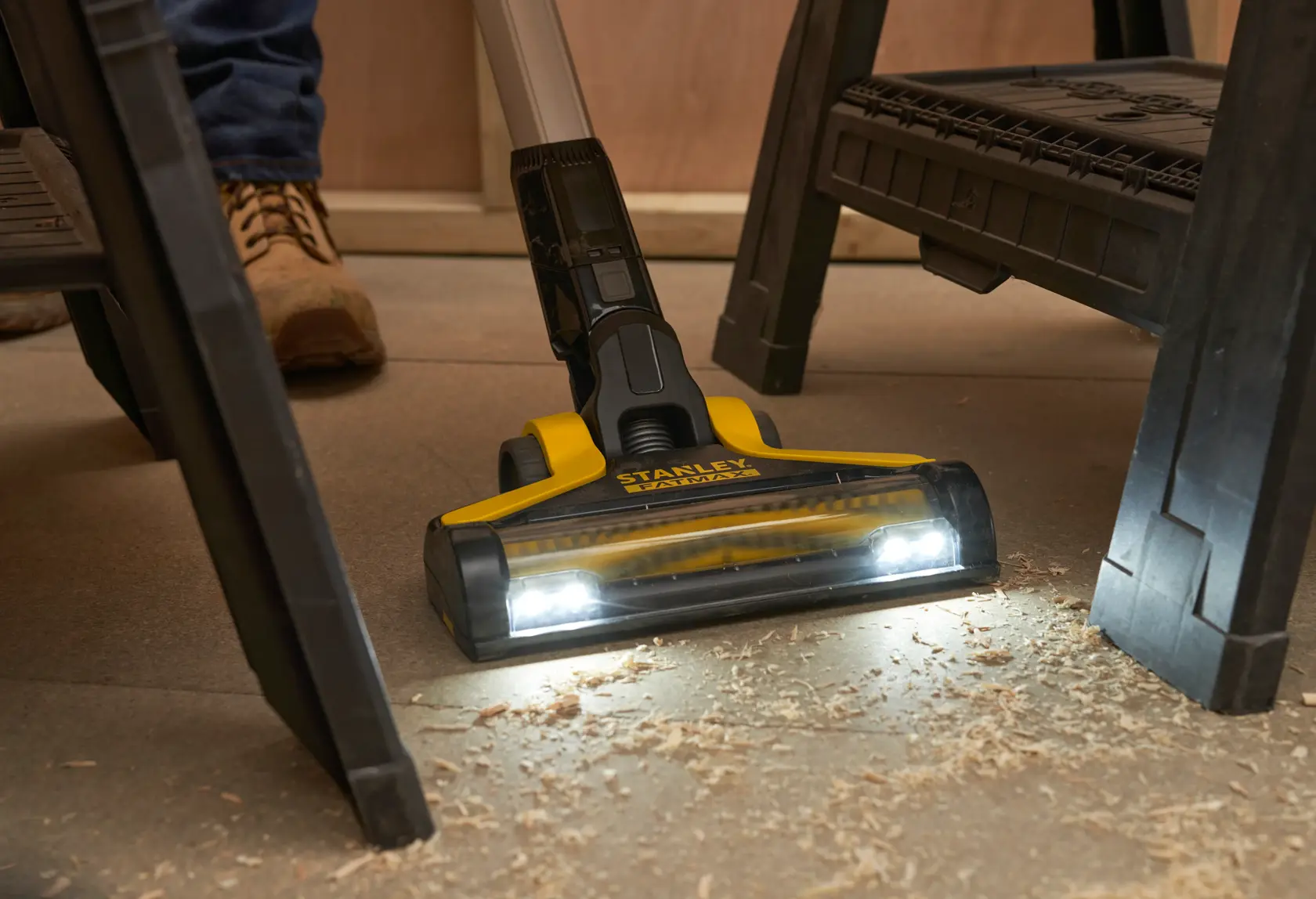 STANLEY SFMCVS001D1 kabelloser Stielstaubsauger mit LED-Leuchten reinigt Holzspäne unter einer Werkbank, Frontansicht.
