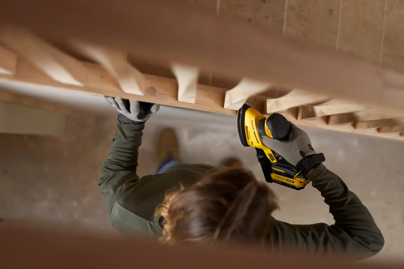 Photo of person using STANLEY SFMCW221D1S cordless sander on wooden railing, wearing gloves.