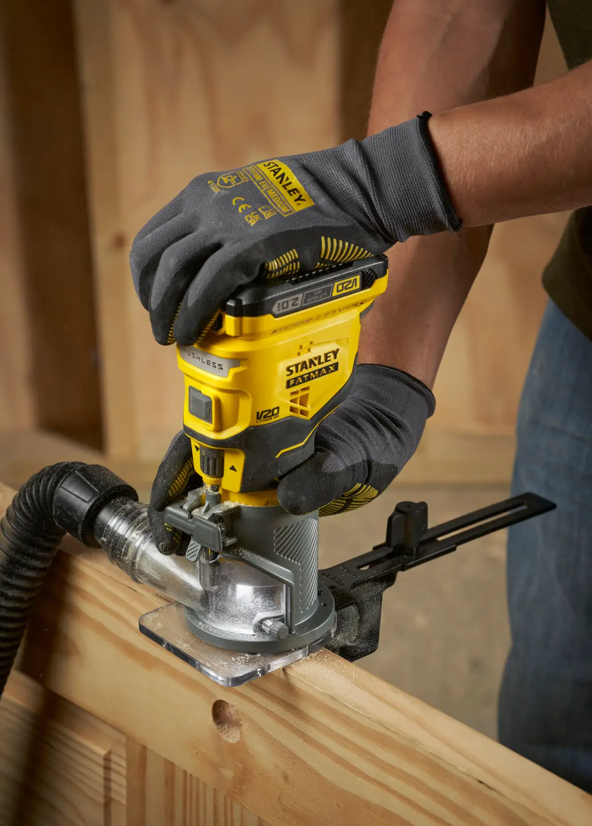 STANLEY SFMCW400B Akku-Oberfräse wird von behandschuhten Händen an Holz verwendet, Nahaufnahme in Werkstatt.