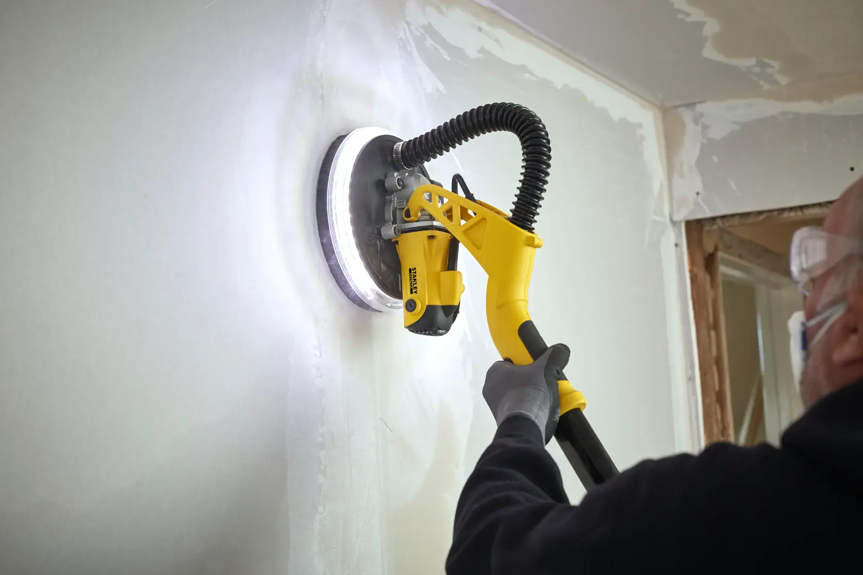 STANLEY SFMEE500S Trockenbauschleifer im Einsatz, gelber Griff, weiße Wand schleifen, Nahaufnahme.