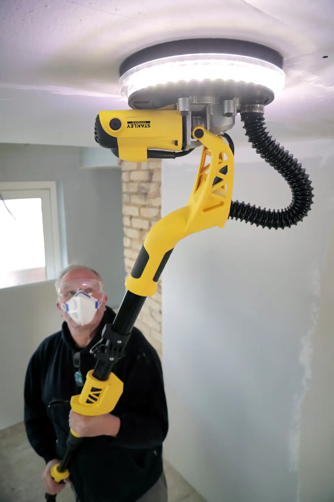 STANLEY SFMEE500S drywall sander in use, yellow and black tool with LED light, sanding a ceiling.