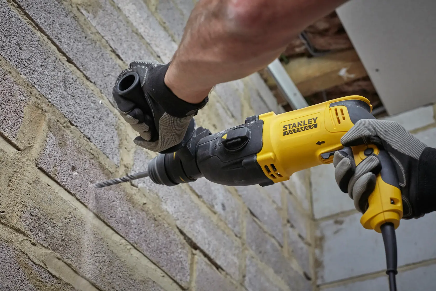 STANLEY SFMEH210K-QS Bohrhammer im Einsatz an einer Ziegelwand, Nahaufnahme von Werkzeug und behandschuhten Händen.