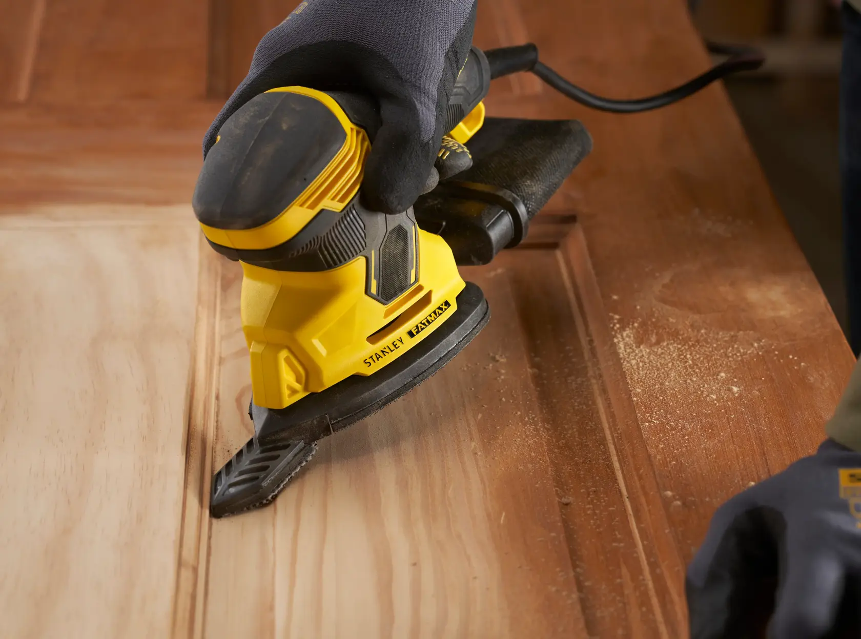 Photo of STANLEY SFMEW210S sander in use on wooden surface, close-up angle showing dust and ergonomic design.