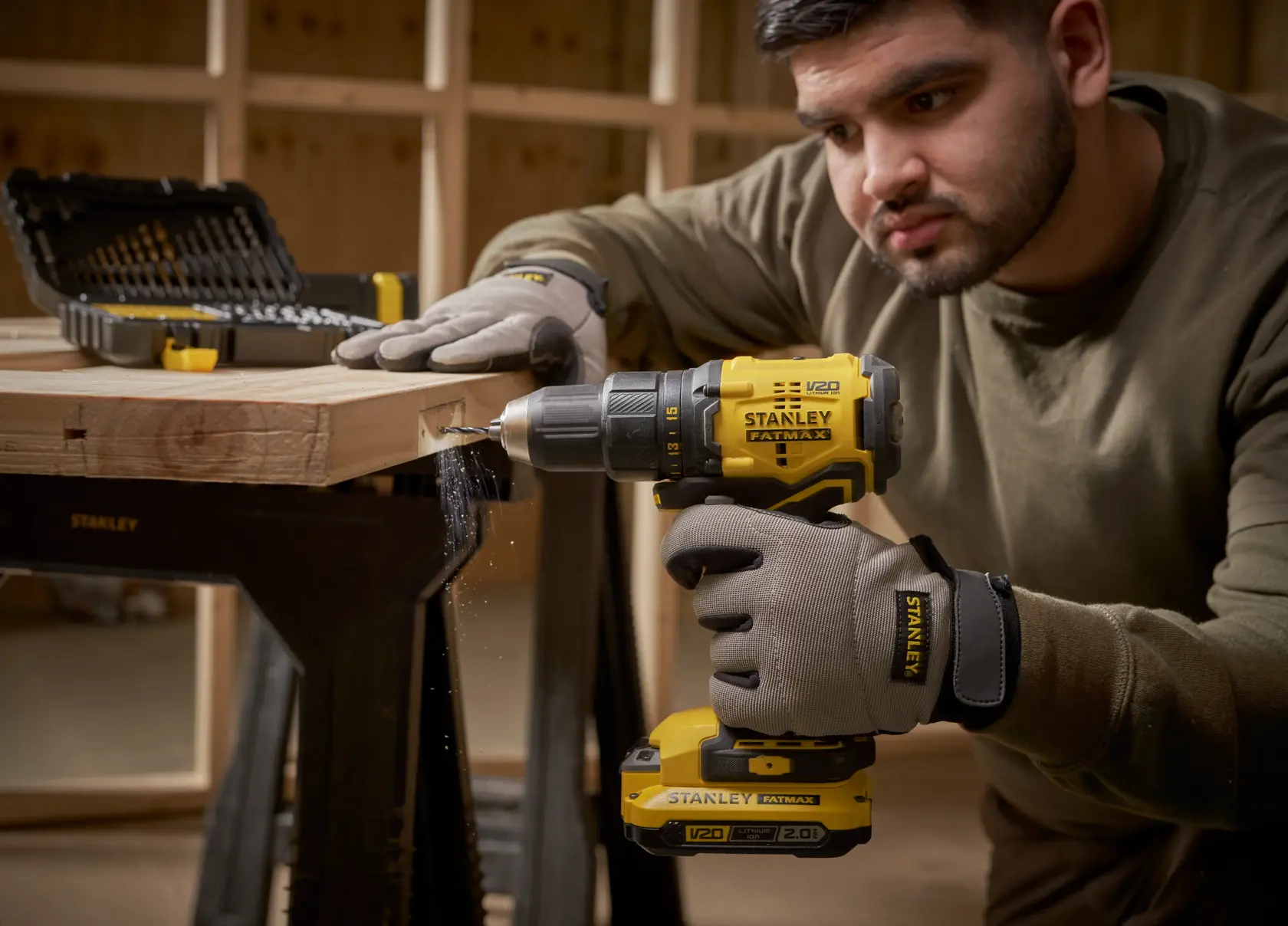 Photo of a STANLEY STA88546 cordless drill in use with gloves, drilling into wood; tool kit visible in background.