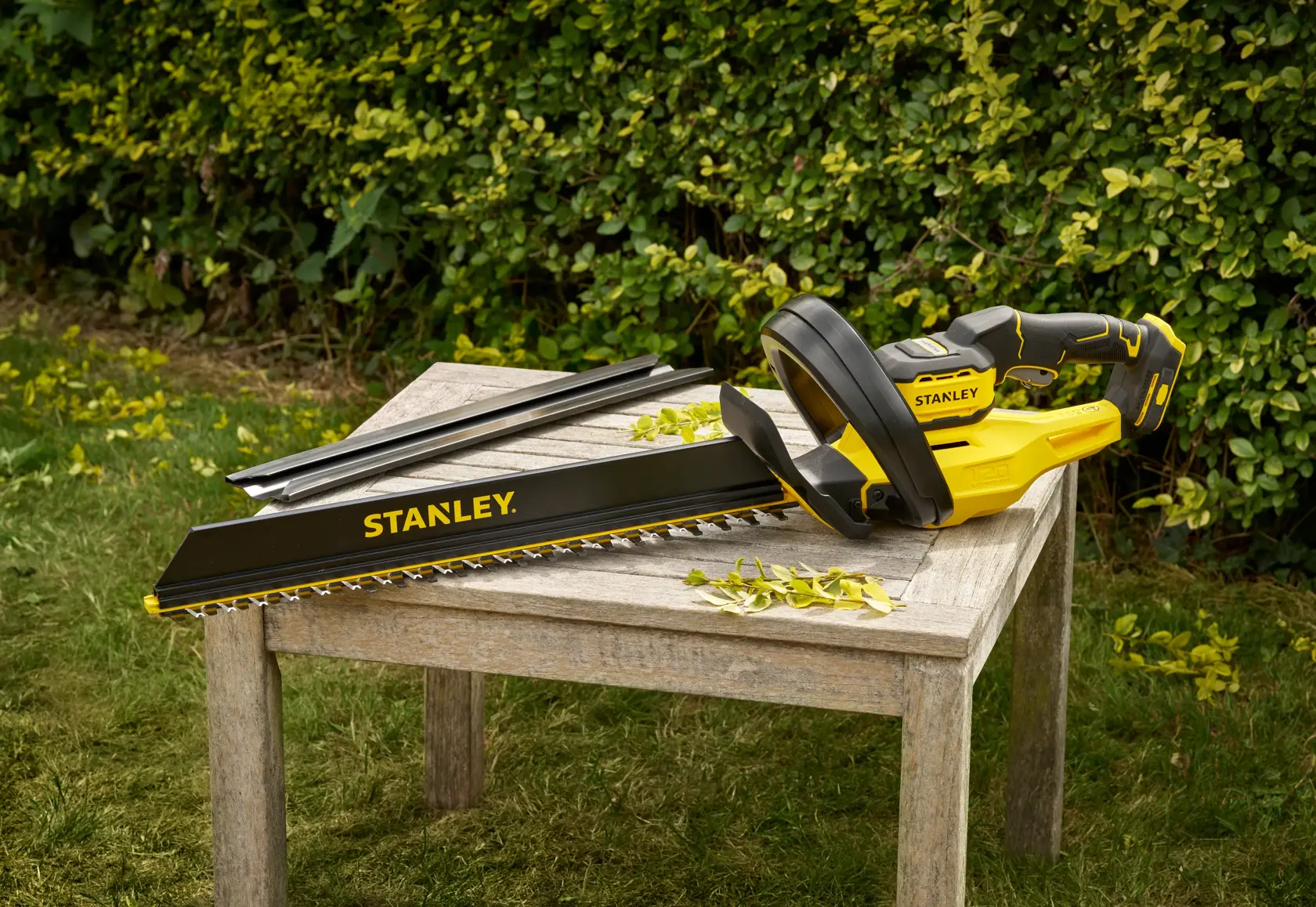 STANLEY STCHT8255B gelb-schwarzer Heckenschneider auf Holztisch im Freien, Klingen und Blätter sichtbar.