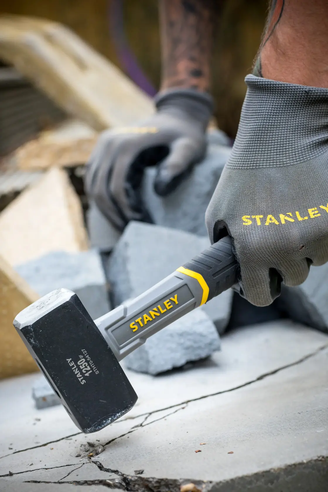 Foto vom STANLEY STHT0-54127 Fäustel im Einsatz, schwarzer Kopf mit gelben Akzenten, von behandschuhter Hand auf Beton gehalten.