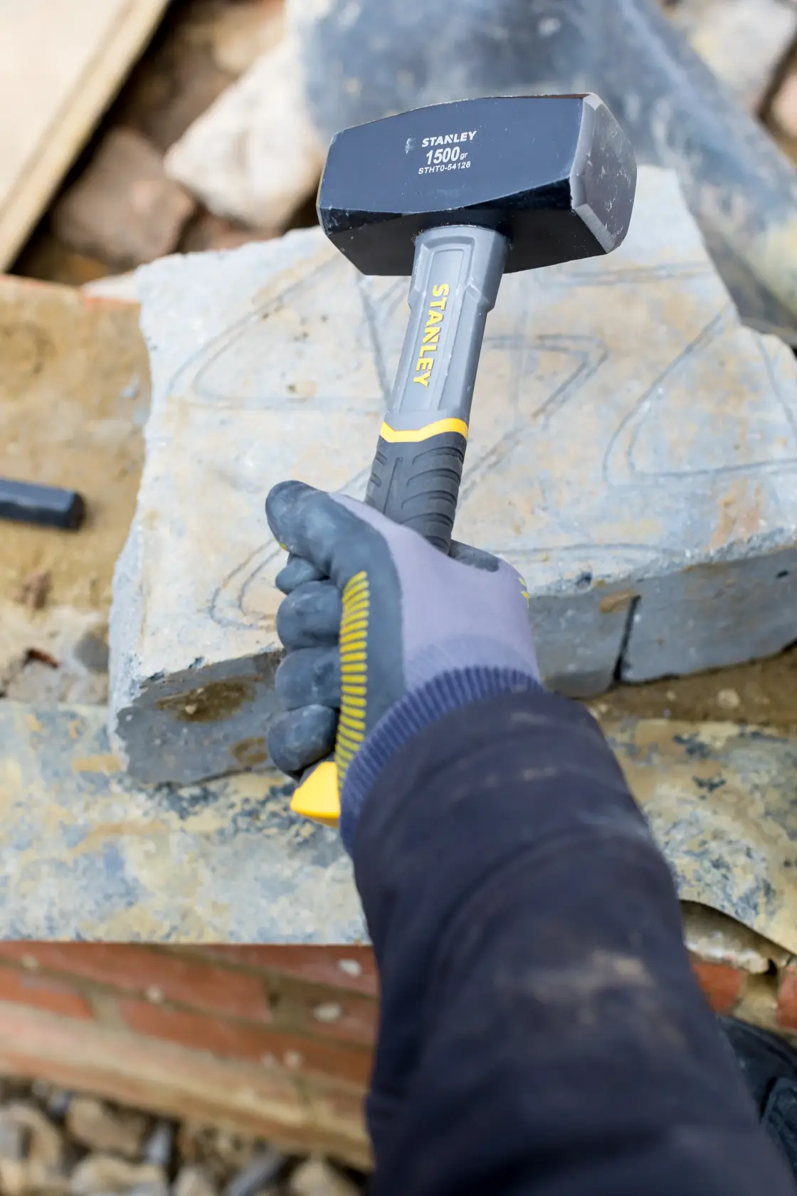 STANLEY STHT0-54128 Fäustel mit gelbem und schwarzem Griff, gehalten von behandschuhter Hand auf Baustelle mit Steinen.