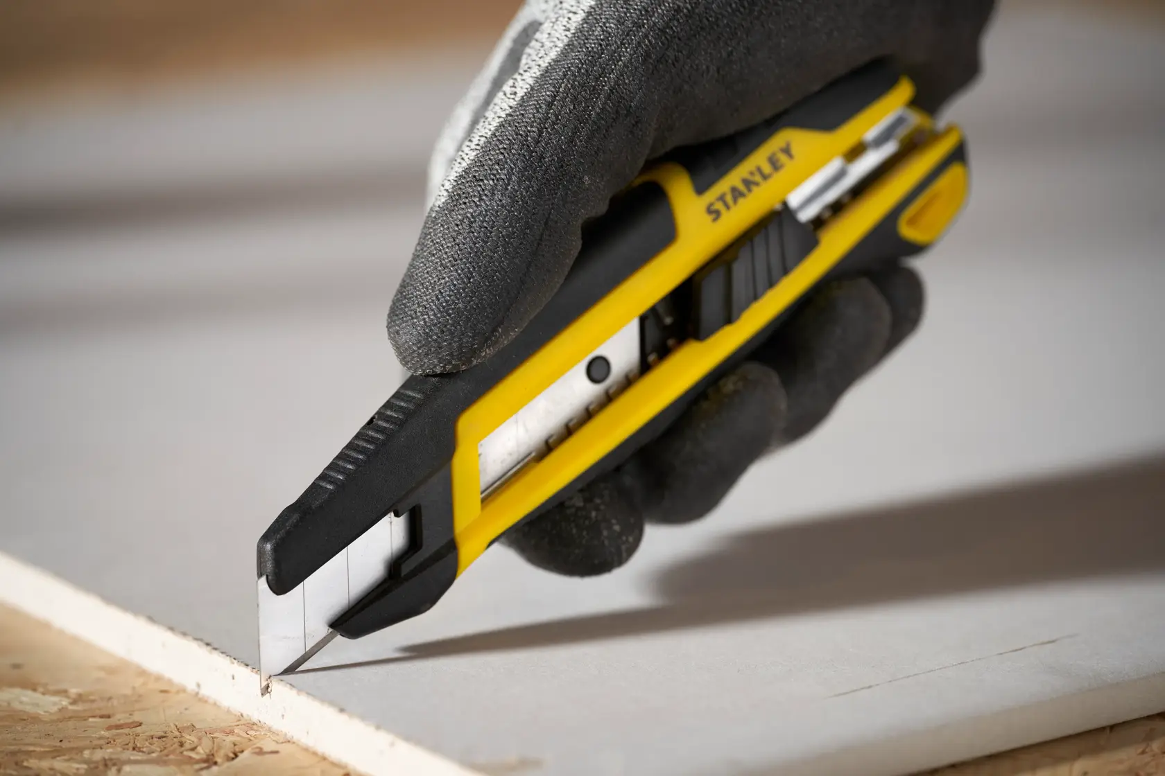 Foto des STANLEY STHT10500-0 Cuttermessers mit gelb-schwarzem Gehäuse, das Gipskarton schneidet, in behandschuhter Hand gehalten.