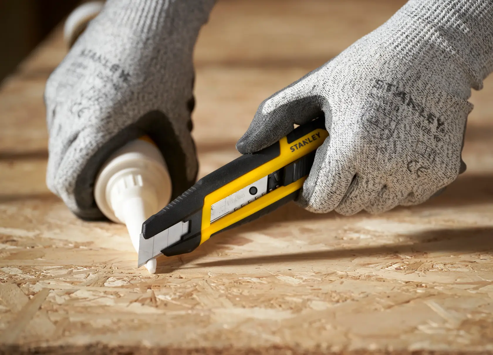 Foto des STANLEY STHT10501-0 Cuttermessers beim Schneiden auf Holzoberfläche, gehalten von behandschuhten Händen.