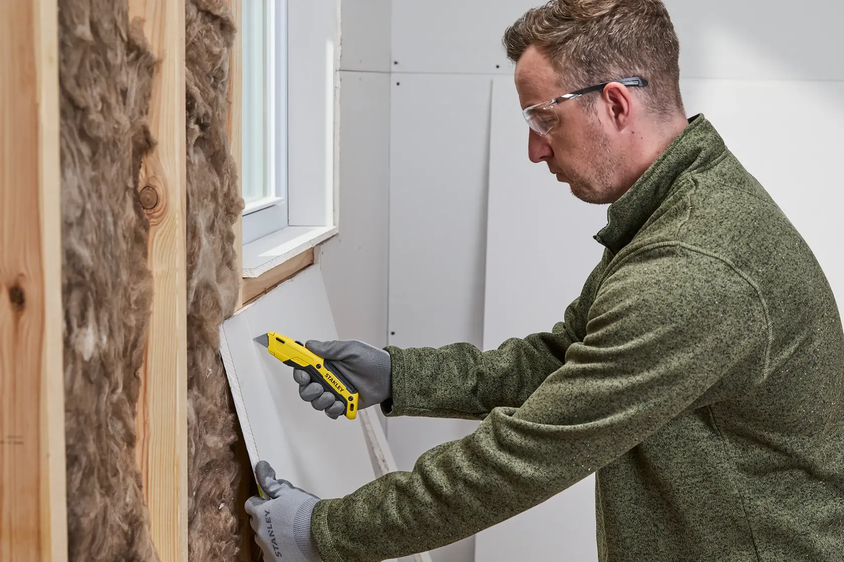 Eine Person mit Handschuhen schneidet mit einem gelben STANLEY Universalmesser ein Stück Trockenbauplatte in der Nähe eines Fensters in einem Gebäude im Bau.