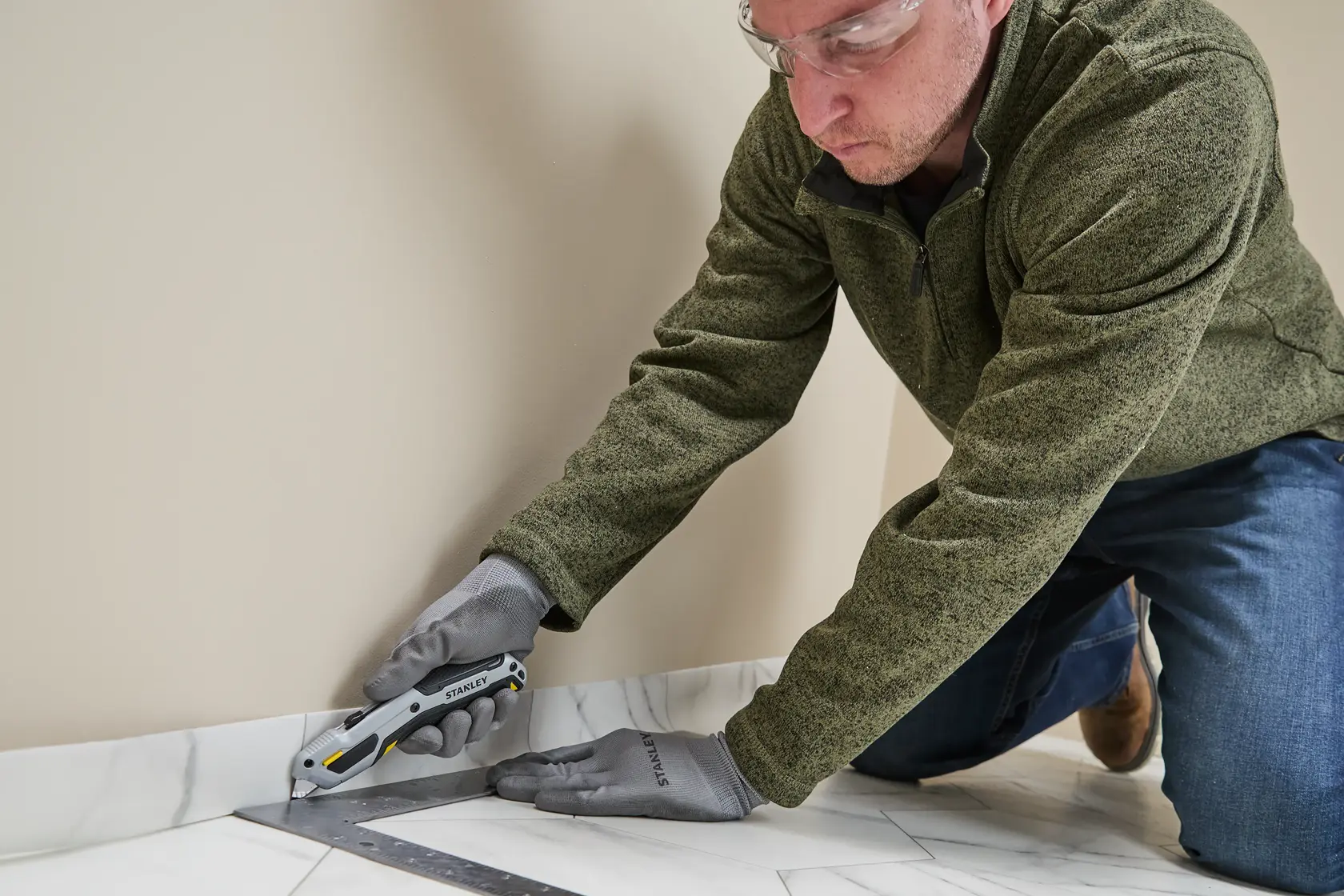 Eine Person mit Handschuhen und grüner Jacke benutzt ein STANLEY Universalmesser (Modell STHT10721-0), um am Rand eines Bodens nahe einer Wand zu schneiden, unterstützt von einem Metallwinkel.