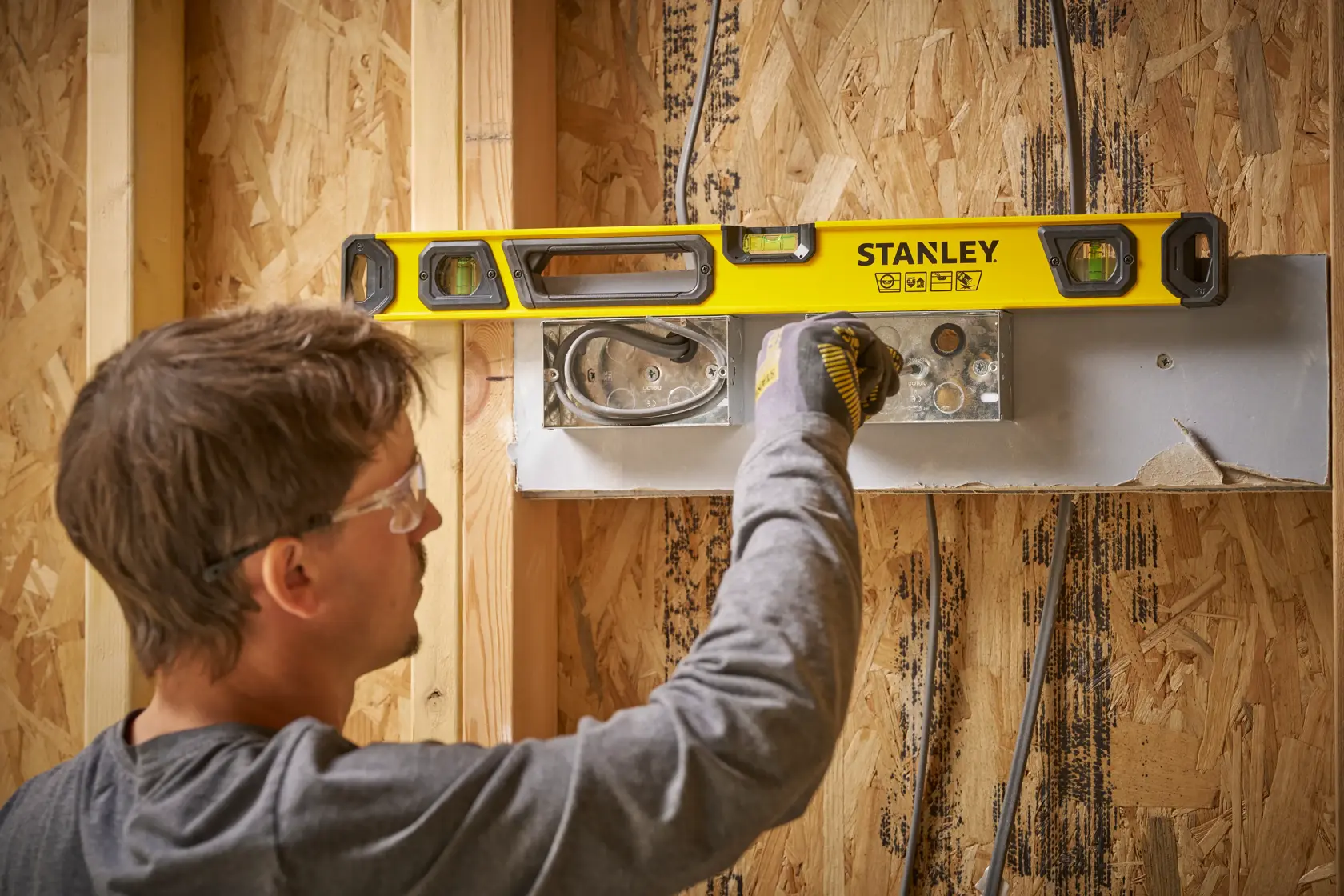 Eine Person verwendet eine gelbe STANLEY Wasserwaage an einem Holzrahmen während der Bauarbeiten. Das Gesicht der Person ist aus Datenschutzgründen unkenntlich gemacht. Die Wasserwaage wird benutzt, um eine Elektroinstallation auszurichten.
