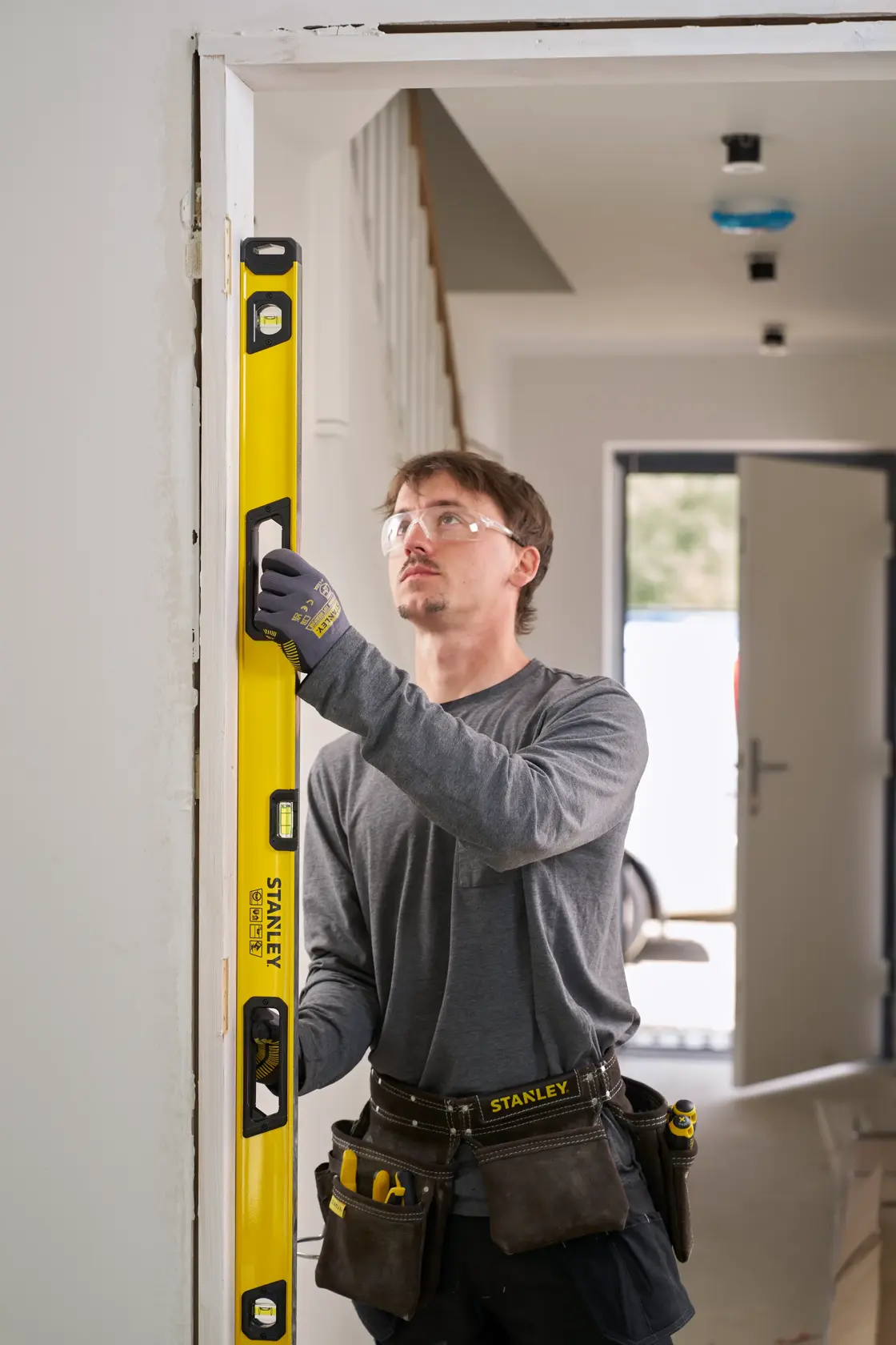 Person benutzt eine gelbe STANLEY Wasserwaage, um die vertikale Ausrichtung eines Türrahmens zu messen. Die Person trägt Arbeitshandschuhe, einen Werkzeuggürtel und legere Arbeitskleidung in einer Innenumgebung.