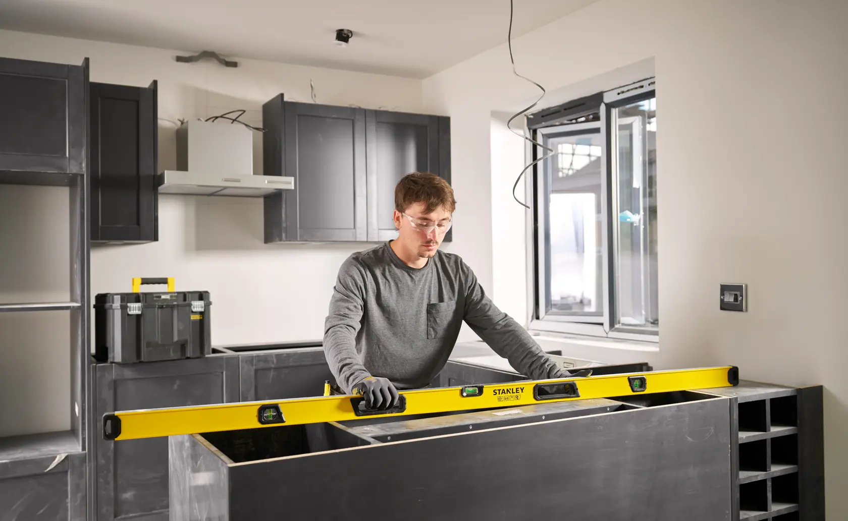 Eine Person verwendet eine gelbe STANLEY Wasserwaage, um die Ausrichtung einer Arbeitsplatte in einer modernen Küche im Bau zu überprüfen. Im Hintergrund sind schwarze Schränke und ein Werkzeugkasten zu sehen. Das Gesicht der Person ist verschwommen.