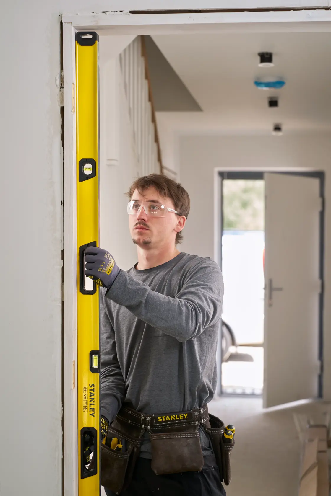 Eine Person mit Arbeitshandschuhen und Werkzeuggürtel verwendet eine gelbe STANLEY Wasserwaage, um die vertikale Ausrichtung eines Türrahmens in einem Haus im Bau oder in Renovierung zu prüfen.