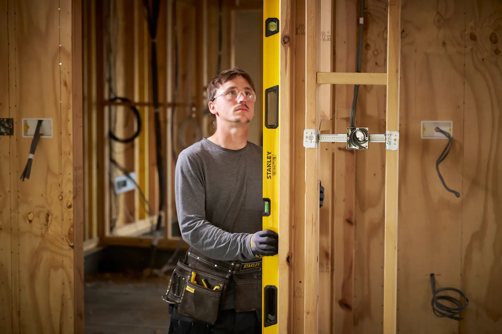 Eine Person mit Arbeitshandschuhen und einem Werkzeuggürtel verwendet eine gelbe Stanley-Wasserwaage, um die vertikale Ausrichtung eines Holzrahmens auf einer Baustelle zu messen. Im Hintergrund sind unfertige Holzwände und elektrische Leitungen zu sehen.