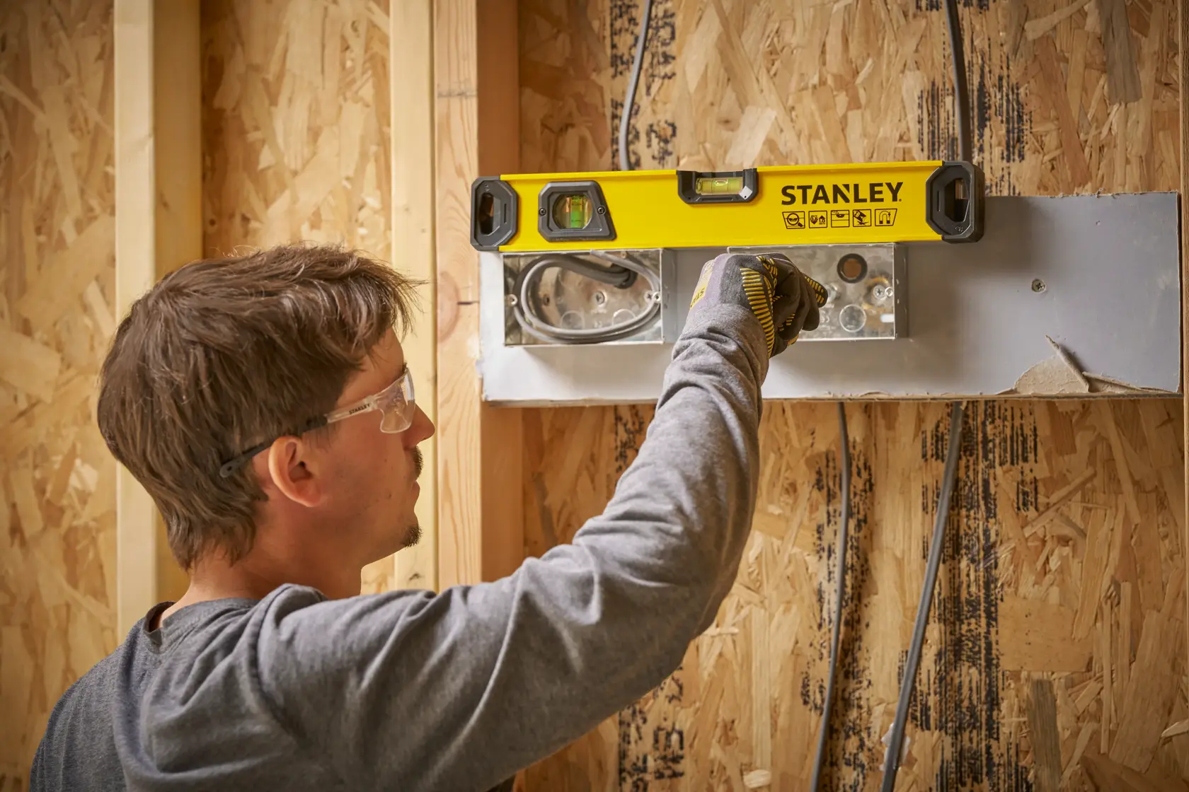 Eine Person verwendet eine STANLEY-Wasserwaage, um die Ausrichtung eines Elektrogehäuses an einer Holzwand während des Baus zu überprüfen. Die Person trägt ein graues Shirt und Arbeitshandschuhe.