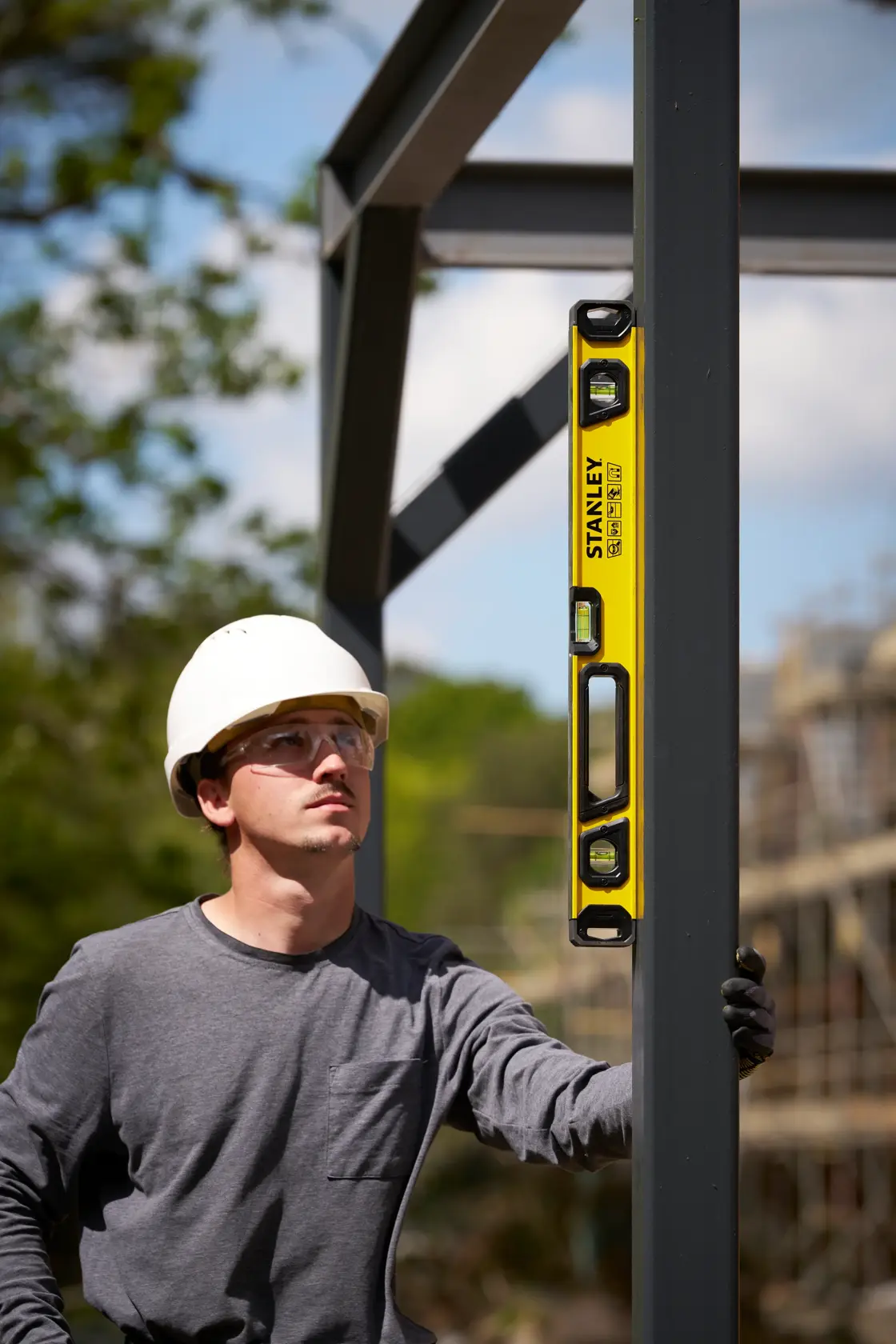 Eine Person mit weißem Schutzhelm überprüft mit einer gelben STANLEY Wasserwaage die Ausrichtung eines vertikalen Metallträgers auf einer Baustelle. Das Gesicht der Person ist verschwommen.